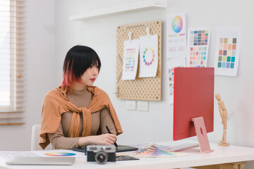 A creative and stylish young female freelance designer is using a tablet and stylus pen while sitting at her desk in her home office.