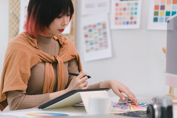 A creative and stylish young female freelance designer is using a tablet and stylus pen while sitting at her desk in office.