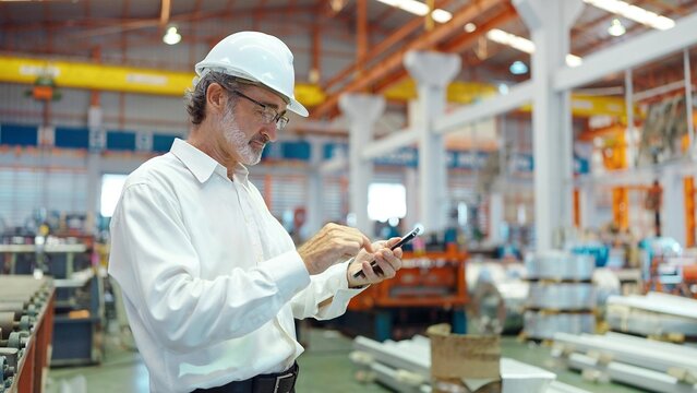 Engineer manager leader man wearing helmet typing on smartphone standing in workplace area at manufacturing factory