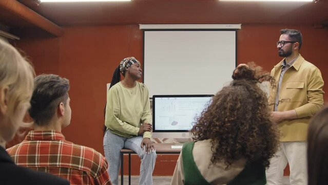 Full arc shot of casually dressed African American female and Middle Eastern male colleagues presenting their project to group of coworkers in conference room of corporate office