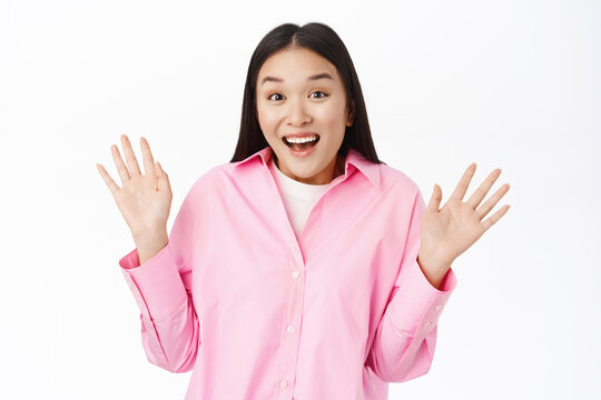Enthusiastic asian girl looks surprised, raise palms, shows empty hands with happy face, stands over white background