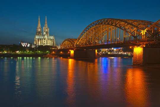 Eee foto in de avond met een lange sluitertijd van de  oude Hohenzollernbrücke over de Rijn in Keulen. Op de achtergod is de mooie verlichte gotische kathedraal de Dom te zien bij een blauwe lucht.