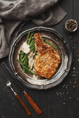 Breaded pork chop with green beans and pepper sauce in vintage plate on dark wooden background. Top view