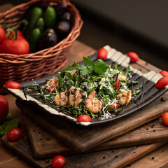 Salad with shrimps, cheese and herbs. Dish on black plate on wooden plank. Wooden table with vegetables. Basket in background. Side view. Soft focus. 