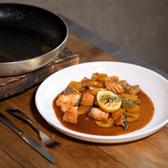Braised salmon with pumpkin in sauce. Dish in white plate with slice of lemon and sprig of rosemary on a wooden table. Gray background. Side view. 