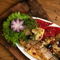 Fried fish with vegetables and herbs. Dish in white plate on wooden plank. Fish with roasted peppers, cauliflower, olives, onions and lettuce. Wooden background. View from above. Close-up.