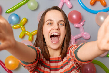 Fototapeta na wymiar Excited woman blogger with brown hair wearing striped dress standing against gray wall decorated with colorful balloons making selfie or broadcasting livestream.