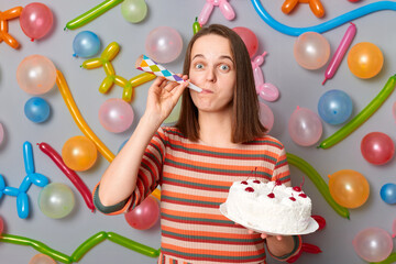 Festive funny happy woman with brown hair wearing striped dress holding cake blowing party horn having fun on party standing against gray wall decorated with colorful balloons.