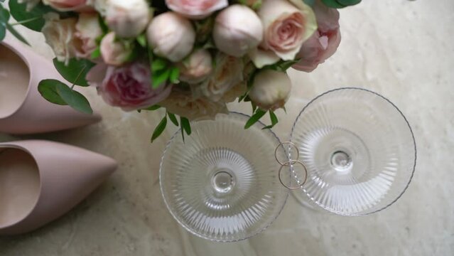 Wedding morning. A bride's bouquet of white and pink peony and roses flowers, women's high-heeled shoes, wedding rings for ceremony and glasses for champagne.