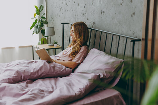 Full body young woman wear purple t-shirt pajama lying in bed read book rest relax spend time in bedroom lounge home in hotel room wake up dream be lost in reverie good mood day. Real estate concept.