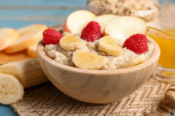 Wooden bowl with tasty oatmeal, raspberries, bananas and apples on blue wooden background