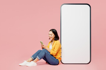 Full body fun young woman of Asian ethnicity wear yellow shirt white t-shirt sit near big huge blank screen mobile cell phone with area use smartphone isolated on plain pastel light pink background.