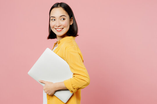 Side View Young Fun Happy IT Woman Of Asian Ethnicity Wear Yellow Shirt White T-shirt Hold Closed Laptop Pc Computer Isolated On Plain Pastel Light Pink Background Studio Portrait. Lifestyle Concept.