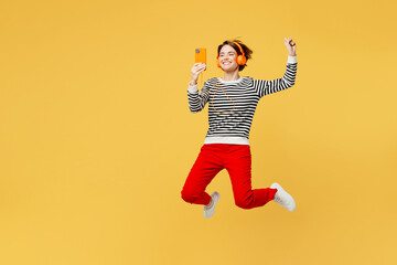 Full body young vivid woman wear casual striped black and white shirt headphones listen music in headphones hold mobile cell phone jump high isolated on plain yellow color background studio portrait.
