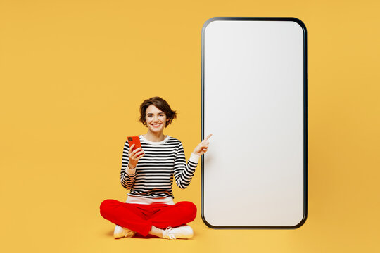 Full body side view young woman wearing casual black and white shirt sit big huge blank screen mobile cell phone with workspace mockup area using smartphone isolated on plain yellow color background.
