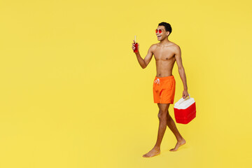 Full body young man wear orange shorts swimsuit glasses hold in hands beer bootle beach refrigerator relax near hotel pool isolated on plain yellow background Summer vacation sea rest sun tan concept