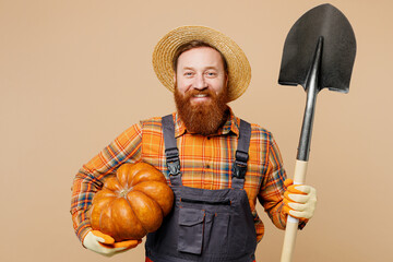 Happy smiling fun young bearded man wear straw hat overalls work in garden hold shovel pumpkin look...