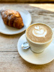 Cup of fresh flat white or latte coffee with croissant on wooden background. French style breakfast in a cafe. Latte art coffee with freshly baked croissant on a wooden table. pov.
