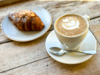 Cup of fresh flat white or latte coffee with croissant on wooden background. French style breakfast in a cafe. Latte art coffee with freshly baked croissant on a wooden table. pov.