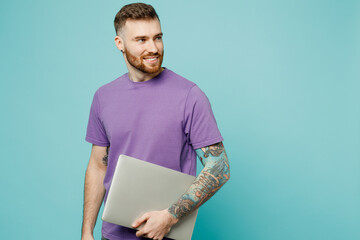 Young smiling fun happy programmer smart IT man he wear purple t-shirt hold closed laptop pc computer look aside isolated on plain pastel light blue cyan background studio portrait. Lifestyle concept.