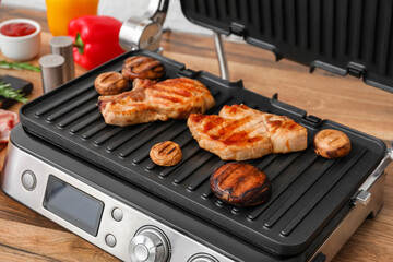 Modern electric grill with tasty steaks and mushrooms on table