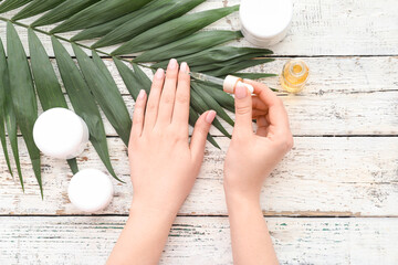 Female hands with bottle of cuticle oil, cosmetics and palm leaf on light wooden background