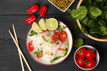 Composition with bowls of tasty rice noodles, vegetables, spinach and lime on black wooden table