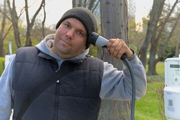 A man holds a cable for charging an electric car connected to a charging station at his head...