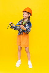 A little girl in a helmet holds a construction tape measure. A child builder in a full-length plaid shirt. The concept of renovation in the children's room. Yellow isolated background.