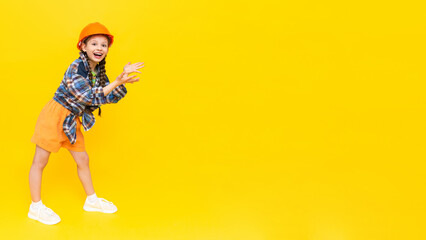 Naklejka premium A child in a construction helmet points to your advertisement. Happy little teen girl in full height. A place to advertise. Yellow isolated background. Copy space. Banner.
