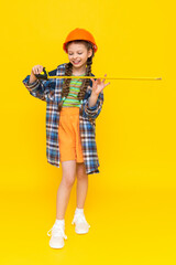 A little girl in a helmet holds a construction tape measure. A child builder in a full-length plaid shirt. The concept of renovation in the children's room. Yellow isolated background.