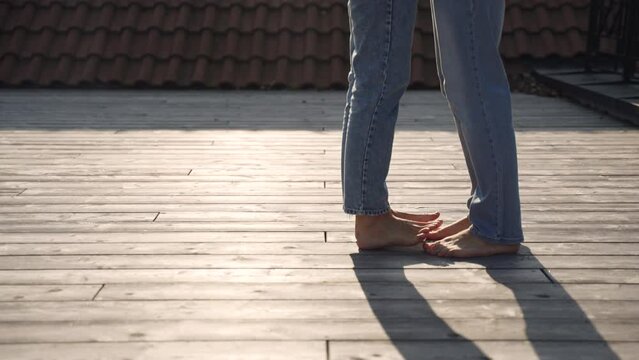 Legs barefoot of woman in jeans on toes playfully approach legs of man and gently step on each other and dance their fingers. Wooden floor, illuminated by sunlight and shadows from couple in love.