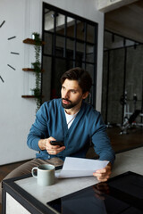 Portrait of worried concerned husband sitting at kitchen counter holding paper document, notification letter and smartphone. Handsome stylish guy dialing number after examining utility bill