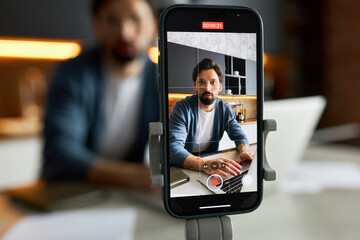 Video blogger influencer recording live on smartphone on tripod, sitting at kitchen table working on laptop, teaching subscribers how to invest and explain financial strategy in trading - obrazy, fototapety, plakaty