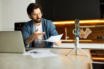 Intelligent man in glasses and business casual clothes sitting at kitchen table with laptop in front of tripod with smartphone, holding paper with financial graph or data explaining trading strategy