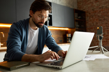 Intelligent smart guy with beard in stylish glasses sitting at kitchen table working on laptop recording video for podcast, typing on keyboard, preparing for live stream. Content creator studying