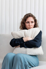 A young woman is sitting on the sofa and hugging a pillow from overflowing sadness during...