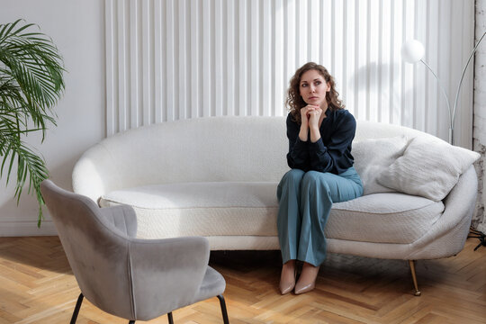 A young woman is sitting on the sofa before a psychological consultation in front of an empty chair. The client is waiting for a specialist in the office