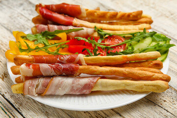 Plate of tasty Italian Grissini with bacon on light wooden background, closeup