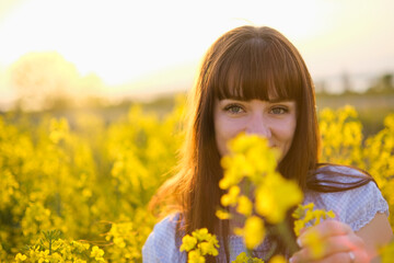 Beautiful girl in a rapeseed field blooming in spring. Holds a branch of a flowering canola in front of him at sunset. The concept of happiness.