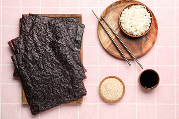 Wooden board with nori sheets, rice, sesame seeds and sauce on pink tile background