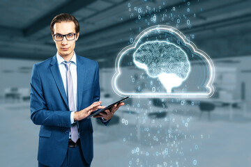 Artificial Intelligence hardware concept. Young businessman with tablet and glowing cloud brain and circuit coding hologram on blurry office background. Data processing, ai trading, machine learning.