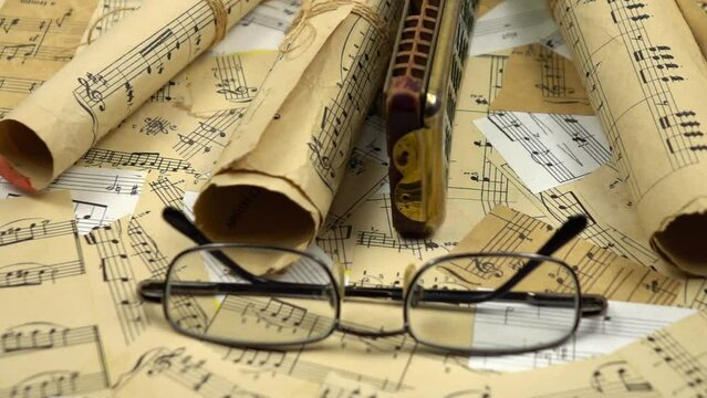 Rolled up sheets music, old harmonica and vintage glasses lie on the various old sheets music, slow motion