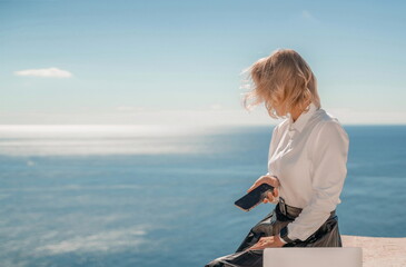 Fototapeta premium Freelance women sea working on a computer. Pretty middle aged woman with computer and phone outdoors with beautiful sea view. The concept of remote work.