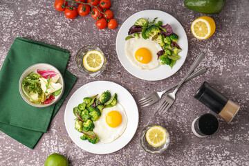 Plates with tasty fried eggs, broccoli and spinach on grey grunge background