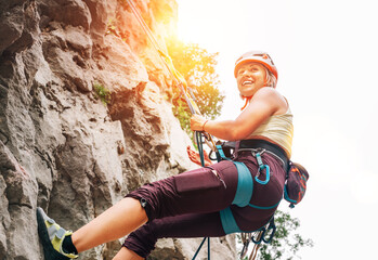 Smiling athletic woman in protective helmet and shoes climbing cliff rock wall using top rope and...