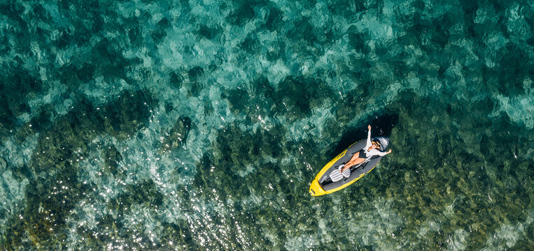 A lonely female in a straw hat smiling, relaxing lying floating in a kayak on the turquoise Adriatic Sea waves. Aerial coastal top view shot. Exotic countries vacations concept.