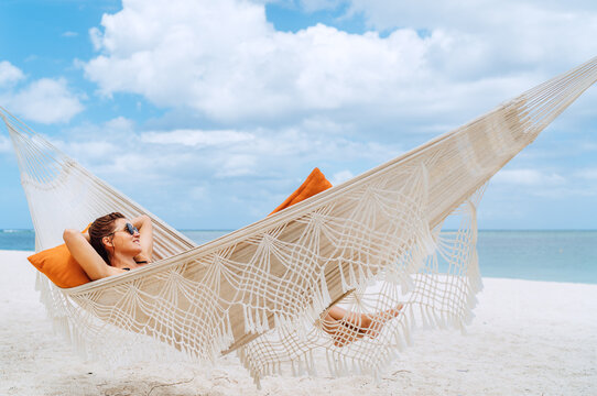 Young woman relaxing in wicker hammock on the sandy beach on Mauritius coast and enjoying wide ocean view waves. Exotic countries vacation and mental health concept image.