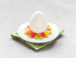 Mousse cream chocolate dessert in the shape of an Easter egg, covered with white mirror glaze. Decorated with colorful candied fruits. On a plate on a linen napkin. Light grey background