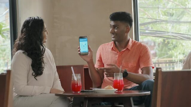 Medium shot of Indian man showing picture on smartphone screen to girlfriend while chatting over drinks in cafe